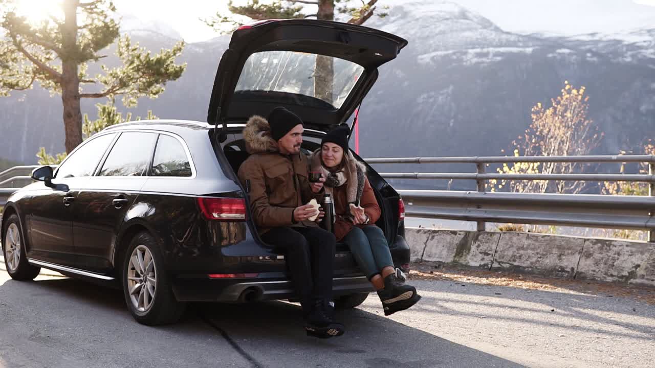 влюбленная молодая пара пьет горячий чай из термоса, сидя в багажнике автомобиля. счастливая кавказская пара делает перерыв на кофе во время автопутешествия по сельской местности - пьет и ест бутерброды. любуется природой. замедленная съемка