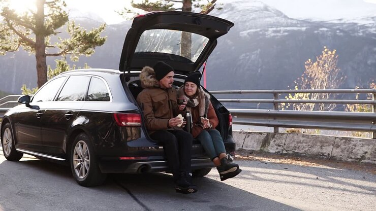Loving, young couple drinks hot tea from thermos flask sitting in car trunk. Happy caucasian couple having a coffee break during road trip in countryside - drink and eating sandwiches. Admiring the nature. Slow motion