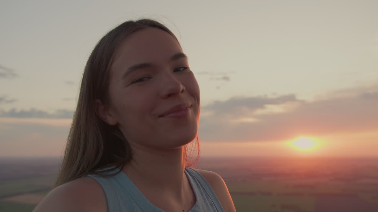 close up profile of young female passenger smiling at camera while gazing at horizon from hot air balloon basket during glowing sunset over vast fields under warm golden light