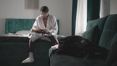 man in bathrobe sitting on bed, reading book in quiet, cozy room. morning light filters through curtains. casual indoor setting with backpack nearby, relaxed and peaceful atmosphere