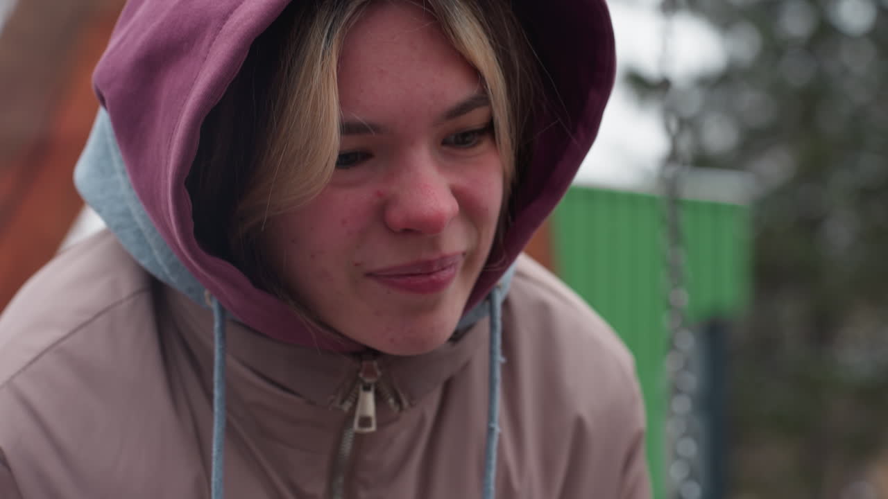 primer plano de una joven inclinada hacia adelante con traje de invierno, sintiendo incomodidad, chaqueta con capucha cubre la cabeza, rostro ligeramente tensado, con fondo borroso con estructuras verdes y elementos al aire libre
