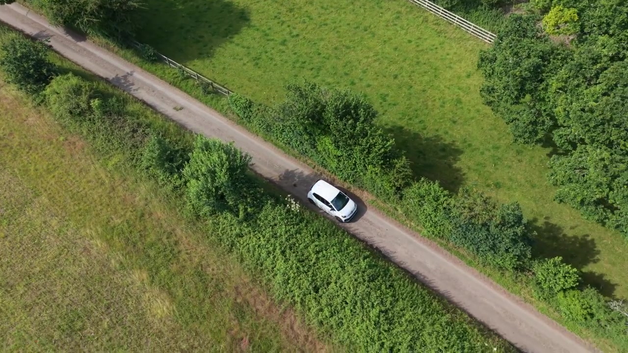 Parco Nazionale South Downs, Hampshire, Inghilterra, filmato aereo con drone di una VW Up! bianca che viaggia lungo una strada di campagna tra campi verdi all'altezza delle cime degli alberi mentre passa sotto gli alberi sotto un sole splendente