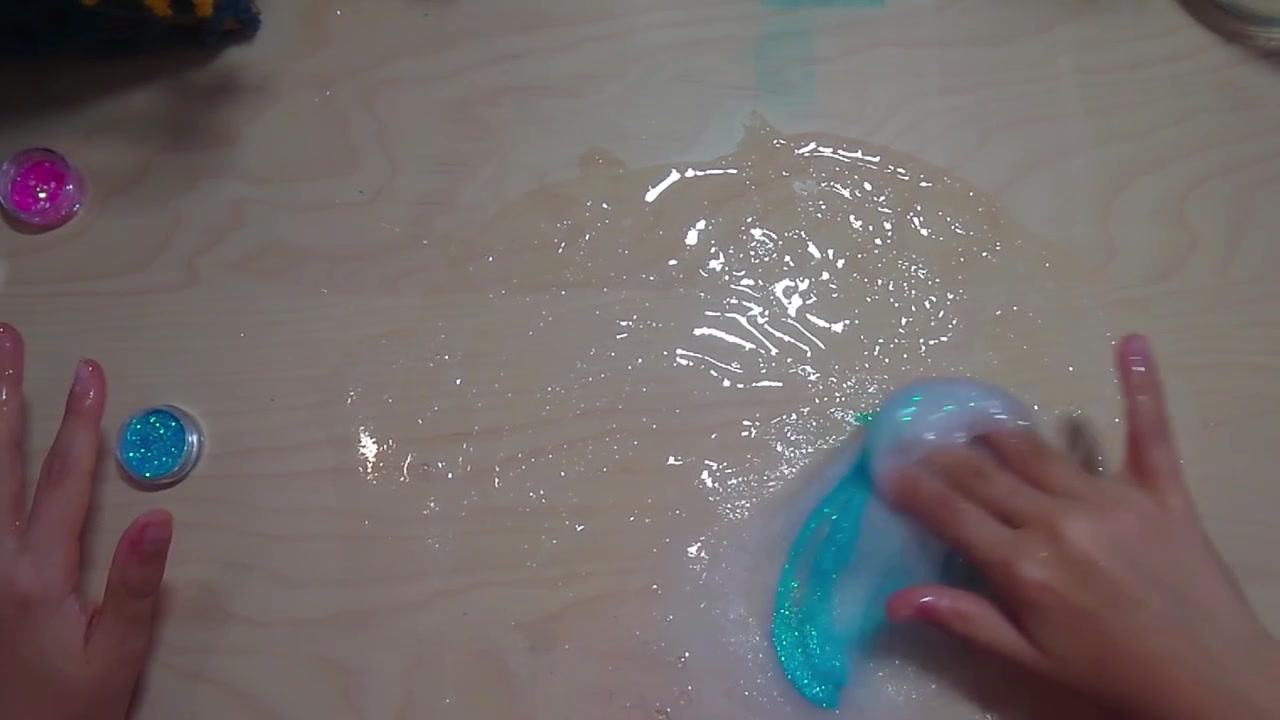 A top-down overhead shot shows a preteen child reaching for a tub of blue glitter while kneading a clear PVA slime base across a wooden desk surface during a fun STEM chemistry craft project.