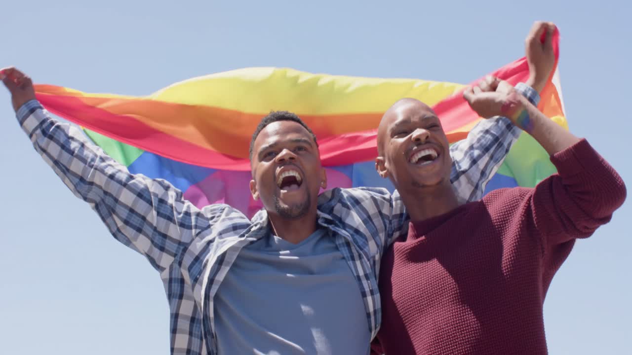 Happy african american gay male couple holding lgbt rainbow flag, slow motion