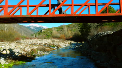 Vista-Aérea-De-Un-Hombre-Caminando-En-Un-Puente-Rojo-De-Montaña-Contemplando-El-Paisaje-De-Los-Pirineos-Españoles