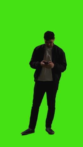 vertical video full length shot of young man talking on mobile phone standing against green screen with low key lighting
