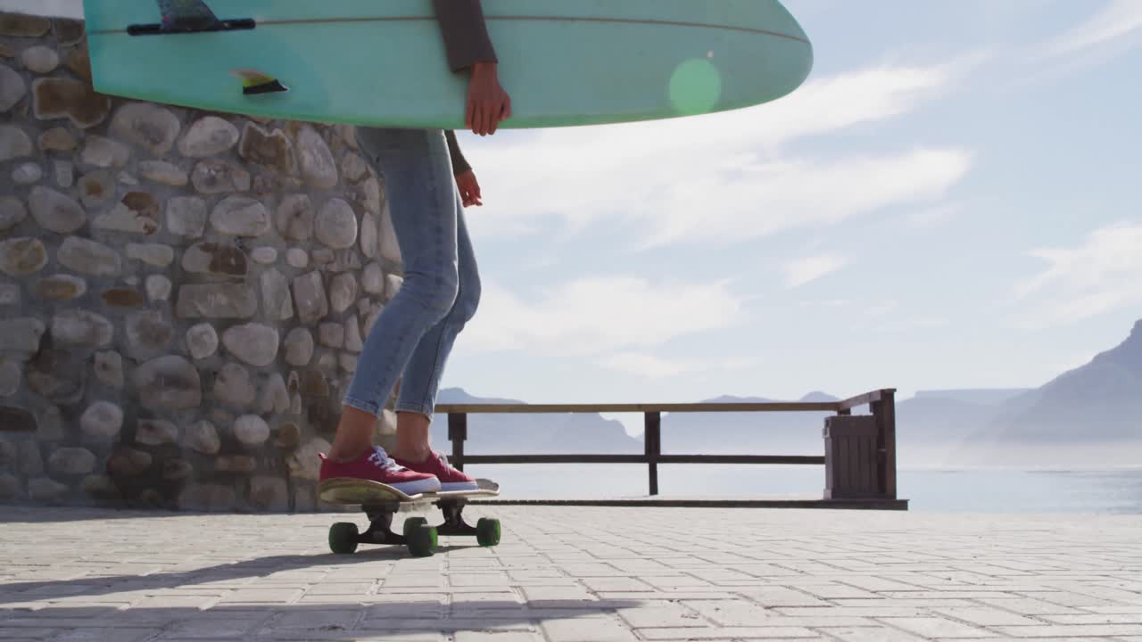 feliz mujer de raza mixta patinando llevando tabla de surf en el soleado paseo marítimo por el mar