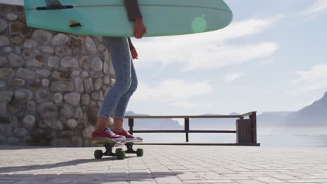 feliz mujer de raza mixta patinando llevando tabla de surf en el soleado paseo marítimo por el mar