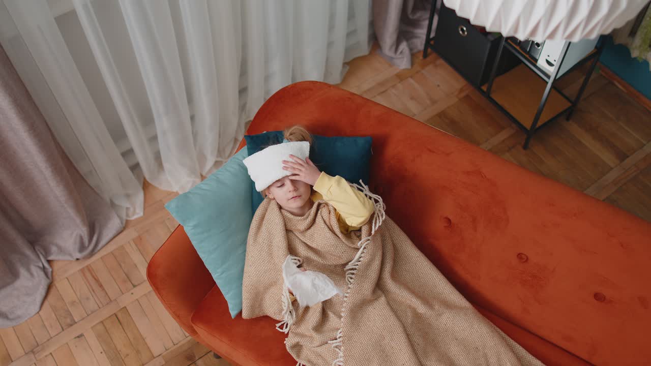 Child girl in plaid suffering from cold or allergy blows nose snot into napkin on sofa at home