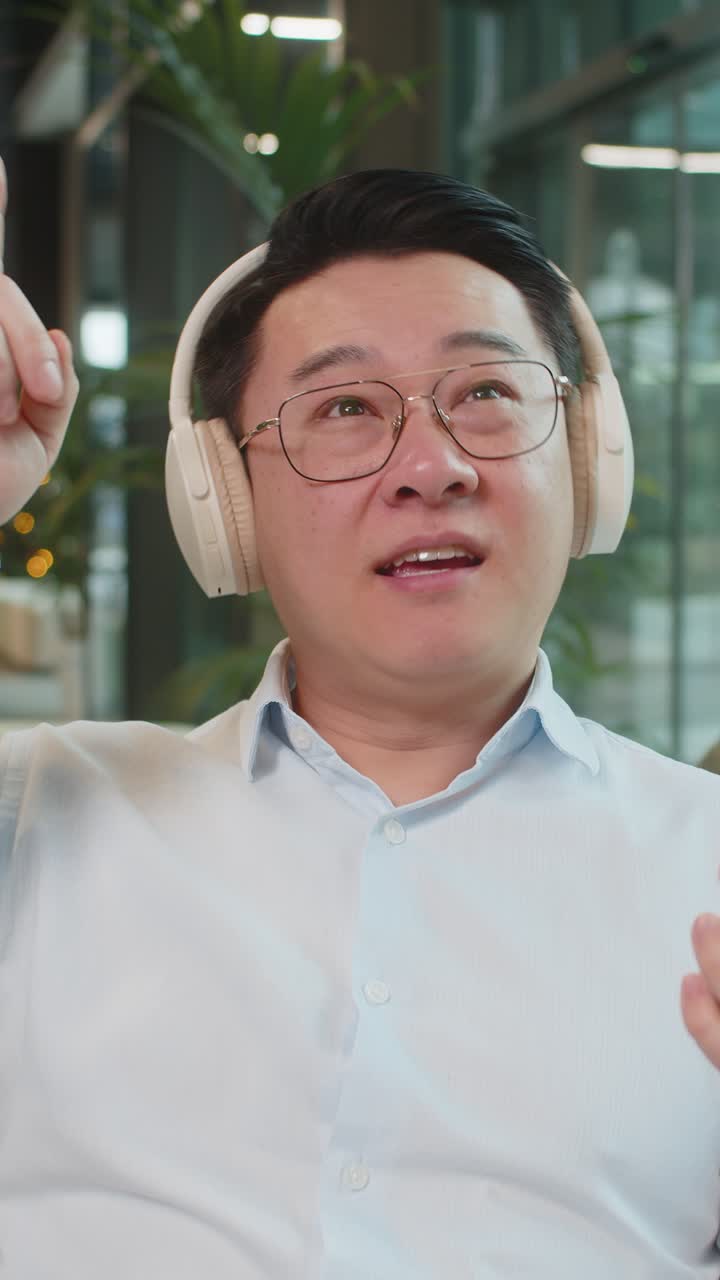 Uomo d'affari maturo eccitato con le cuffie che ascolta musica energetica sullo smartphone nell'atrio dell'ufficio