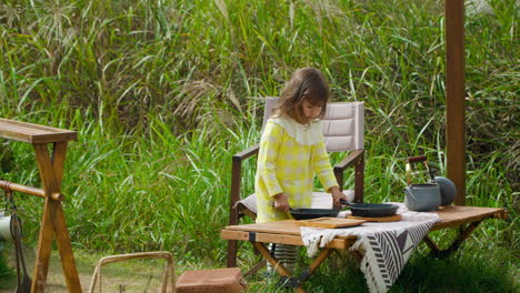 curious little girl playing with frying pans standing by wooden table at camping ground outdoors in country