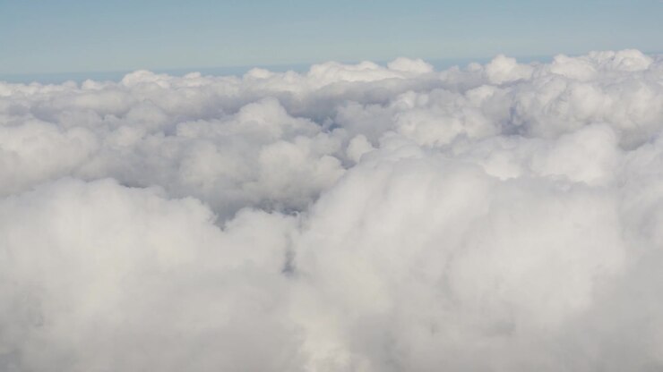 Clouds are visible from above, displaying different shapes and densities. The view shows layers of white clouds against a clear blue sky. The shifting patterns create a dynamic landscape.