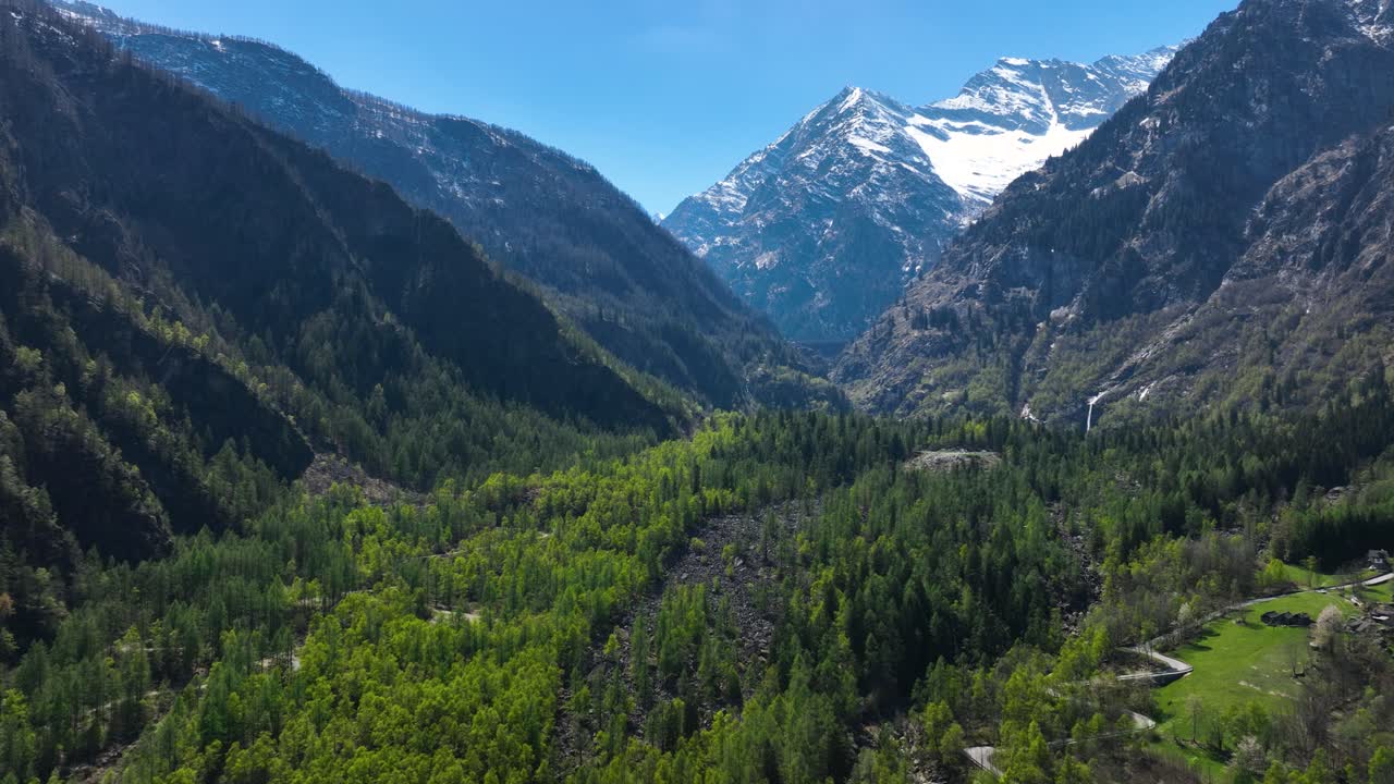 toma aérea lenta de una hermosa montaña cubierta de nieve que se eleva sobre un valle y un bosque con un camino sinuoso y cascadas