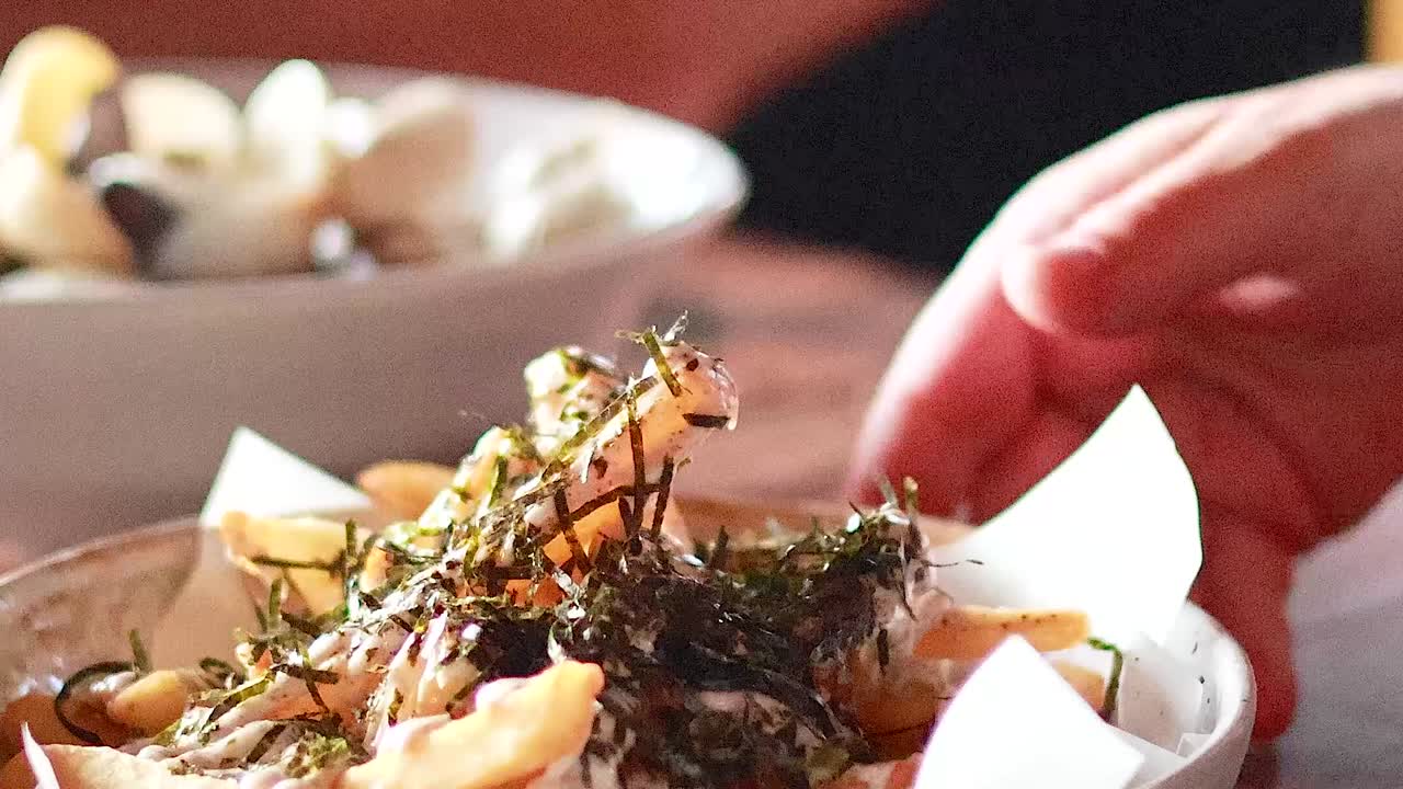 Close-up of hands adding herbs to a dish of fries and cheese on a wooden table.