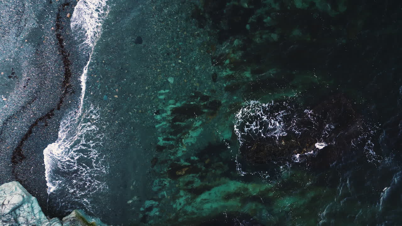 Ocean waves swirl in shallow grey beach with pronounced tide line of seaweed, drone top down