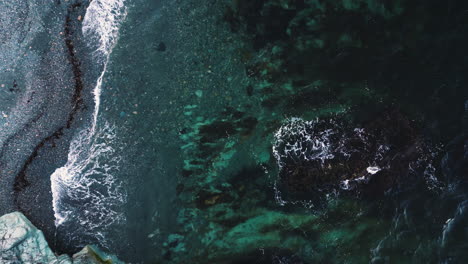 ocean waves swirl in shallow grey beach with pronounced tide line of seaweed, drone top down