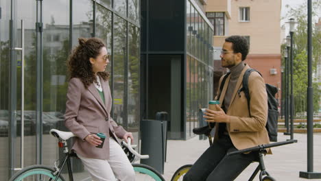 young american man and woman in formal clothes drinking coffee and talking while leaning on their bikes in the street