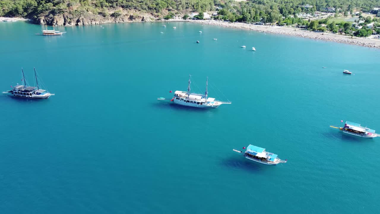 Aerial drone view showing calm turquoise water with anchored boats near sunlit shoreline in Antalya with clear horizon, gentle waves, bright summer colors, peaceful coastal scene