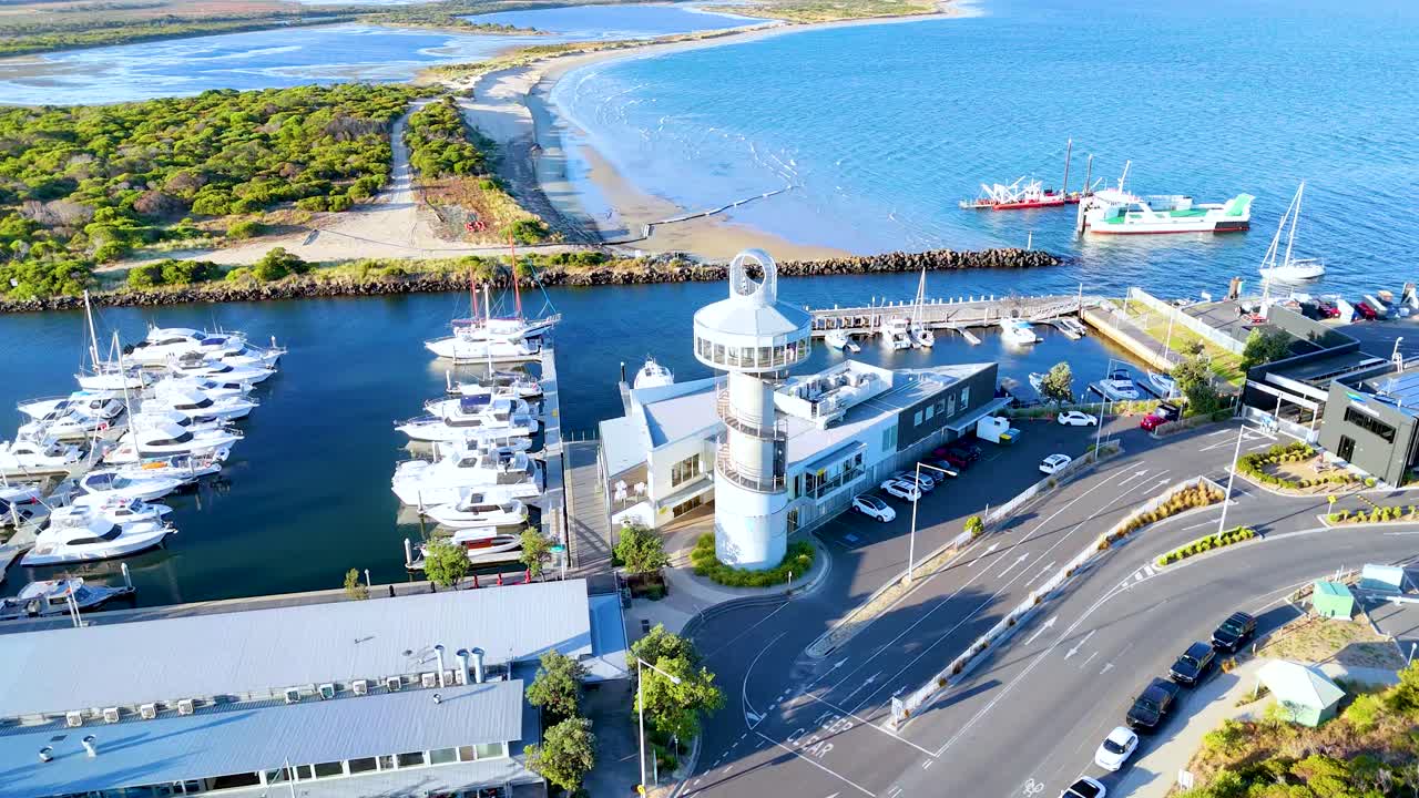 Drone footage captures Queenscliff Marina and lighthouse under bright daylight, showcasing boats, roads, and surrounding landscapes