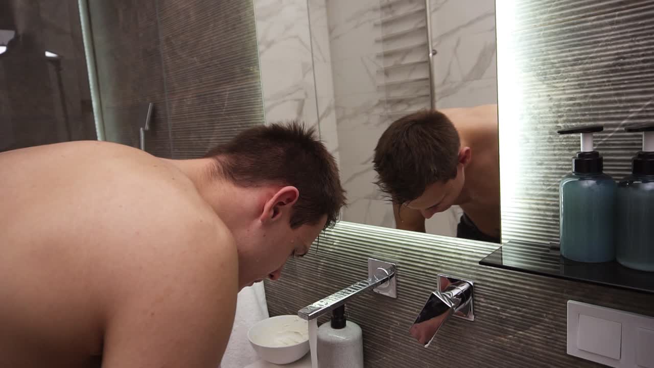 수 ⁇ 에서 차가운 물이 흐르는 싱크대에서 얼굴을  ⁇ 고 있는  ⁇ 벗은 잘생긴 남자. 아침 일상적인 청소 절차를 하는 남자. 거울 반영을 보고