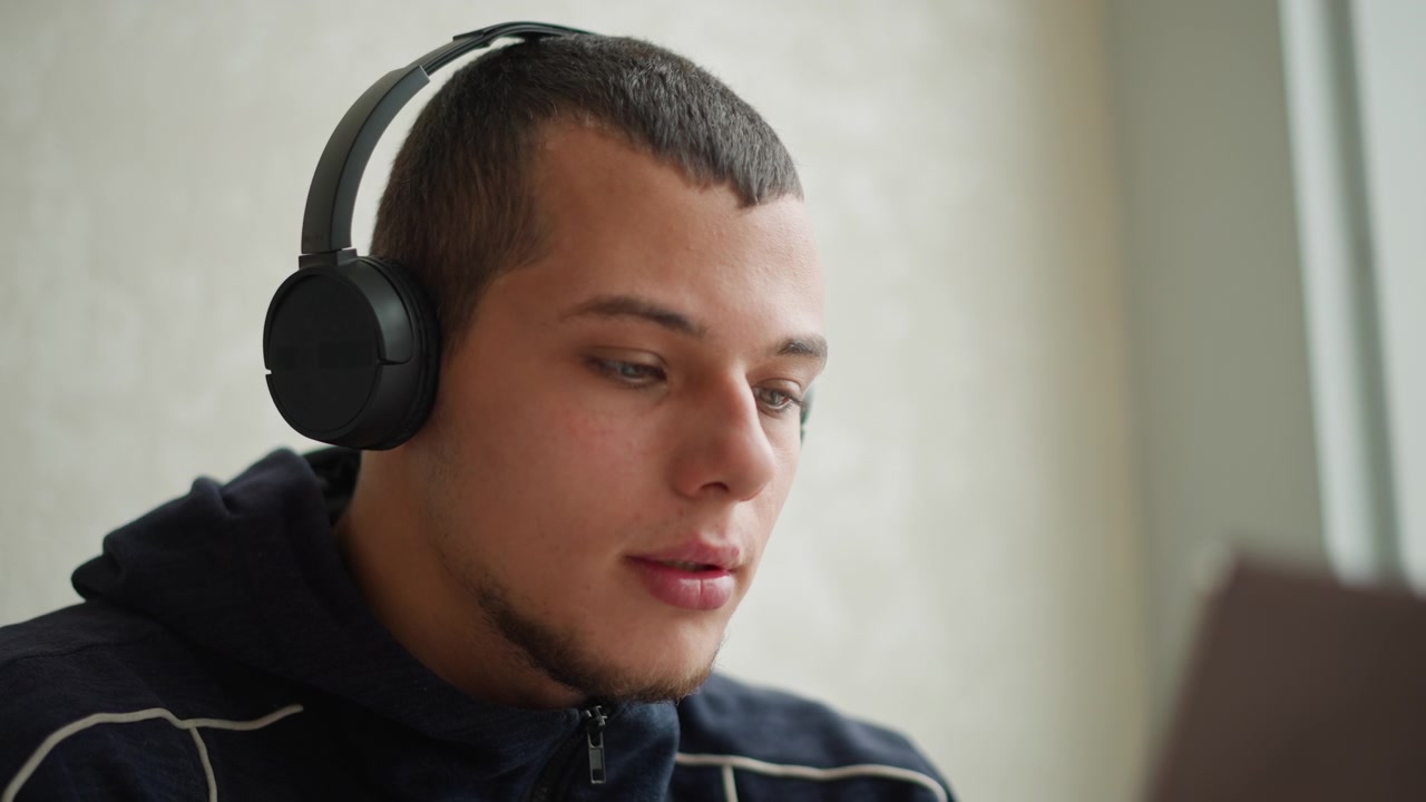 Primo piano di un giovane uomo con cuffie over-ear nere e felpa con cappuccio blu scuro, concentrato sullo schermo con un leggero movimento della testa. L'illuminazione soffusa evidenzia un'espressione calma contro una parete chiara.