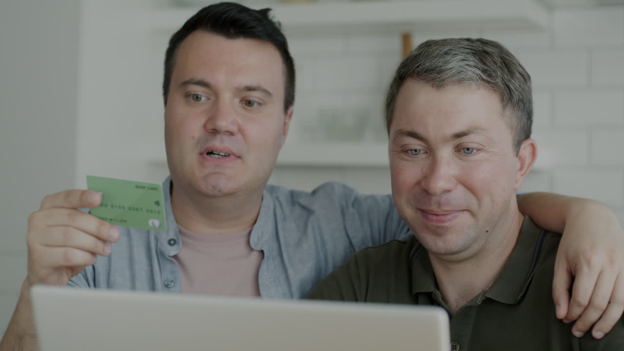 Two men using a laptop and credit card for online shopping at home.