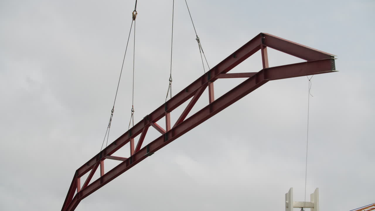 Large Red Metal Truss Being Lifted by Cables Against a Cloudy Sky