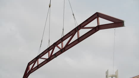 large red metal truss being lifted by cables against a cloudy sky