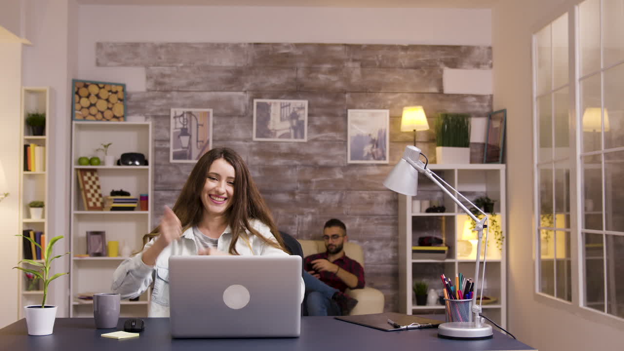A woman working on her laptop while a man sits on the couch in the background