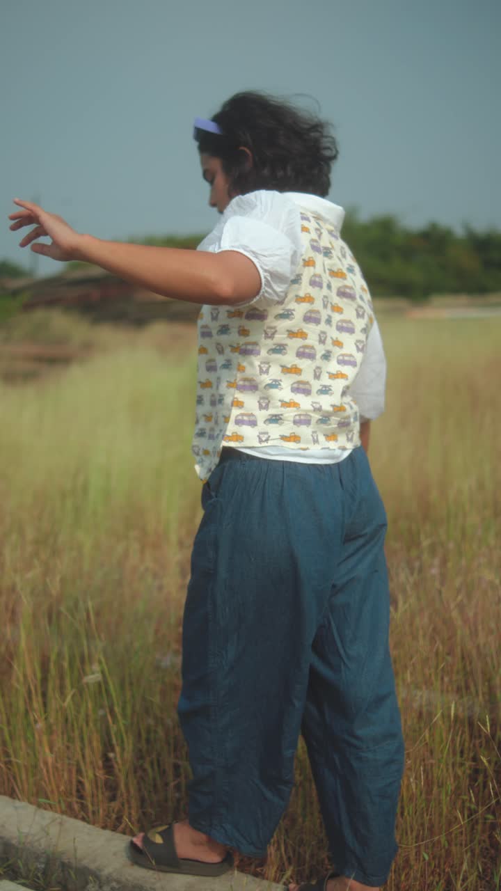 Young woman walking along a concrete block in a grassy field, balancing outdoors in warm daylight. A playful, carefree moment capturing freedom, simplicity, and natural rural energy. Vertical video
