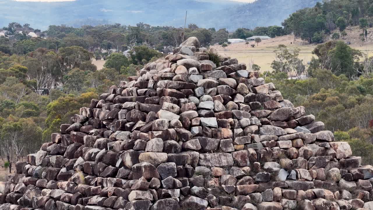 Camera smoothly ascends a large granite rock pyramid in a rural Australian landscape, revealing detailed stone textures and surrounding bushland under soft daylight
