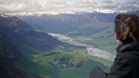 Girl-looking-over-a-beautiful-green-valley-with-mountains-with-a-river-running-through-on-a-bright-sunny-day-on-a-grassy-hill,-drifting-handheld-shot