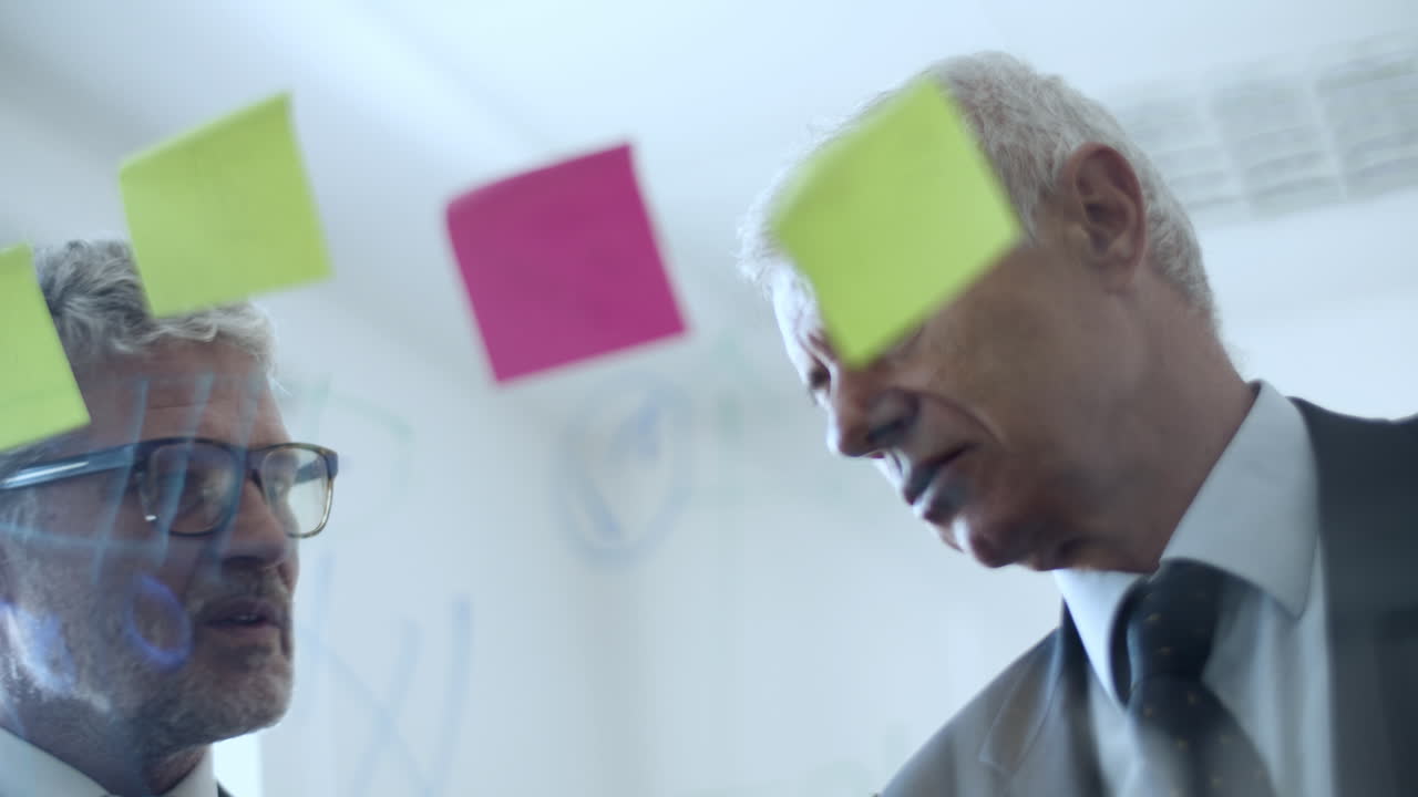 Two serious grey haired male business partners talking in front of sticky notes