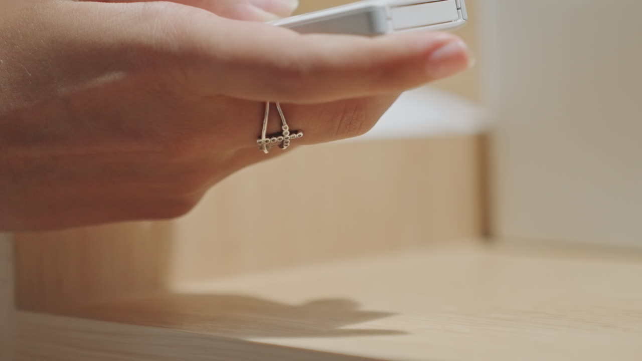 Mano che si allunga verso il telecomando dell'hotel sul tavolo, primo piano della mano di un ospite con anello che solleva un telecomando minimalista da un comodino in legno, luce soffusa della lampada e ombra sottile, gesto di controllo in camera che si mostra
