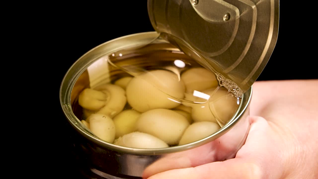 A hand opens a metal can of champignon mushrooms in brine under bright studio lighting, with close-up shots and a steady camera angle