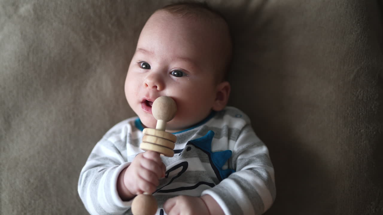 Lovely child holding wooden toy at his mouth. Baby boy trying to chew the toy while lying on his back. Close up.