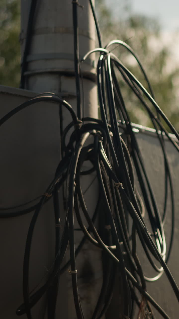 Primer plano de cables enredados en un poste de servicios públicos, detalle urbano cinematográfico con bobinas retroiluminadas y metal erosionado. Fondo bokeh iluminado por el sol, sombras profundas y aislamiento texturizado que crean una atmósfera industrial melancólica.