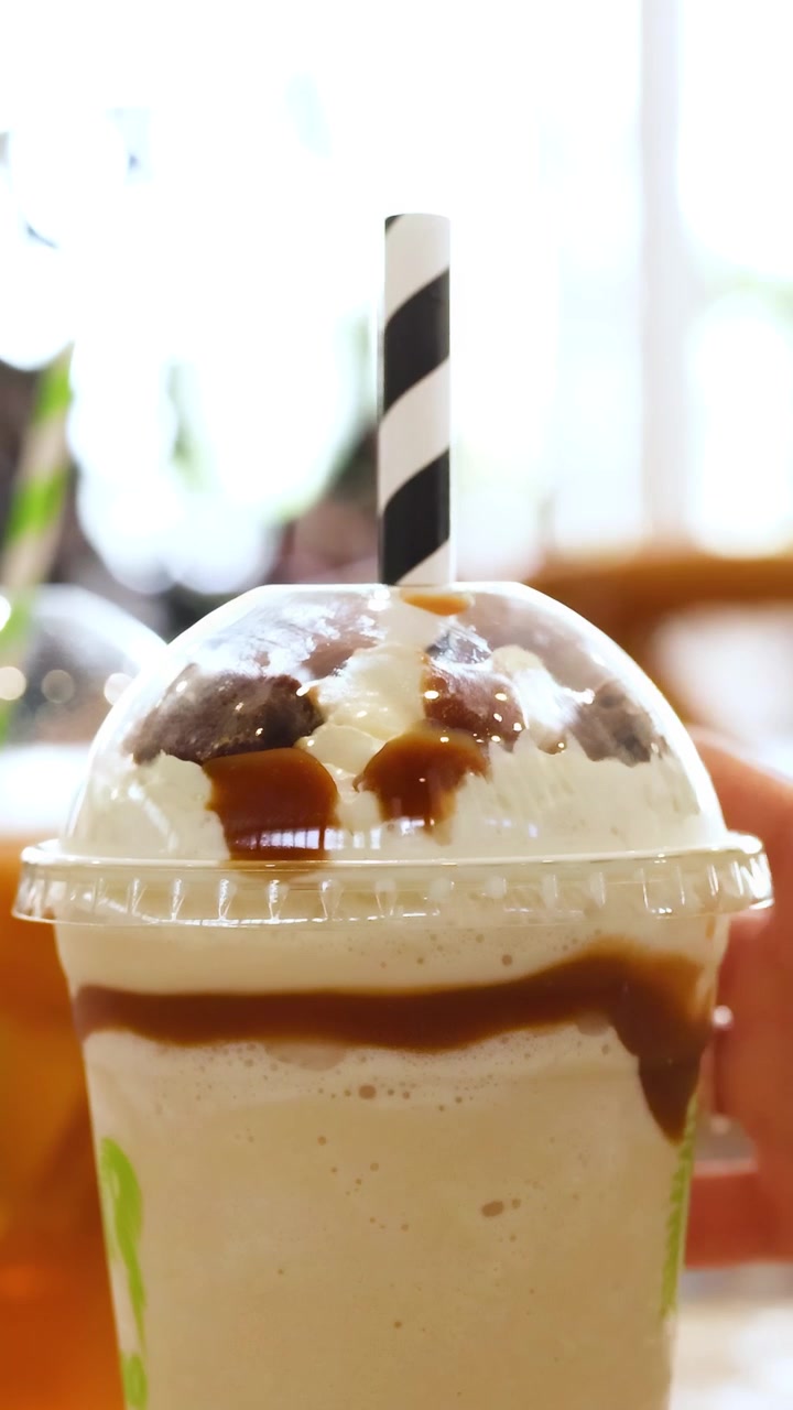 Una mano inserisce una cannuccia a strisce in un milkshake con caramello in un ambiente di caffè ben illuminato.