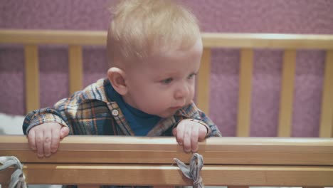 baby-stands-leaning-on-edge-of-wooden-cot-in-light-room