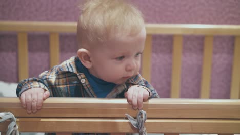 baby stands leaning on edge of wooden cot in light room