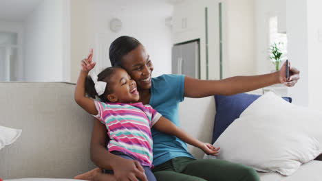 Happy-african-american-mother-and-daughter-sitting-on-sofa-taking-selfie-at-home