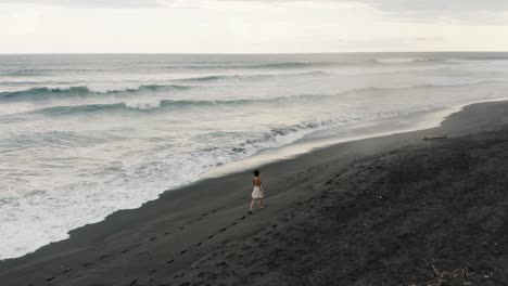 Woman-Walking-On-The-Black-Sand-Beach-Of-El-Paredon-In-Guatemala---aerial-drone-shot