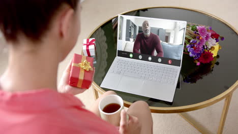 caucasian woman holding red gift using laptop with african american man on screen