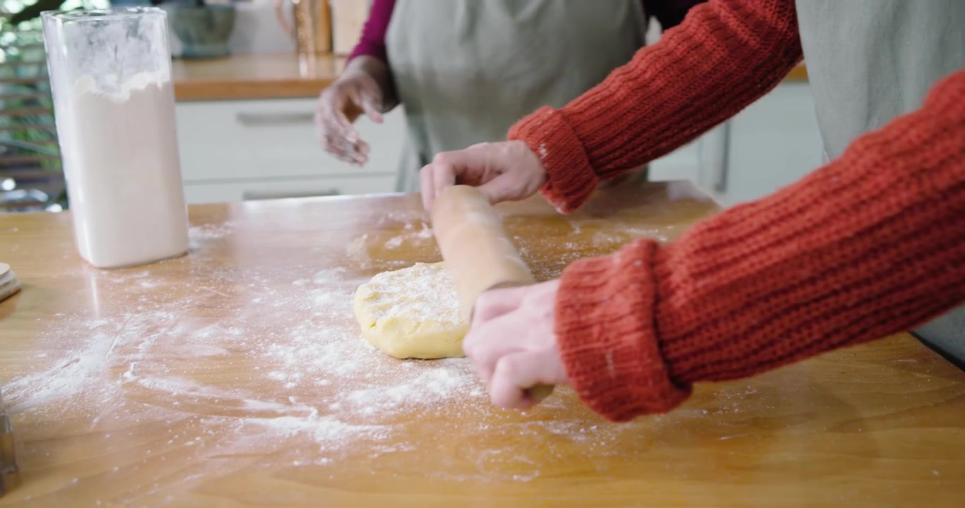 Diverse women friends holding dough in kitchen dusting flour using rolling pin and making cookies
