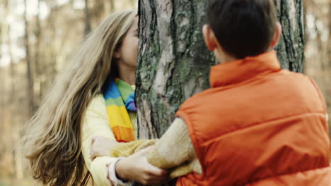 vista de cerca de la hermana y el hermano caucásicos jugando en el bosque, escondiéndose detrás de un tronco de árbol y mirándose desde diferentes lados