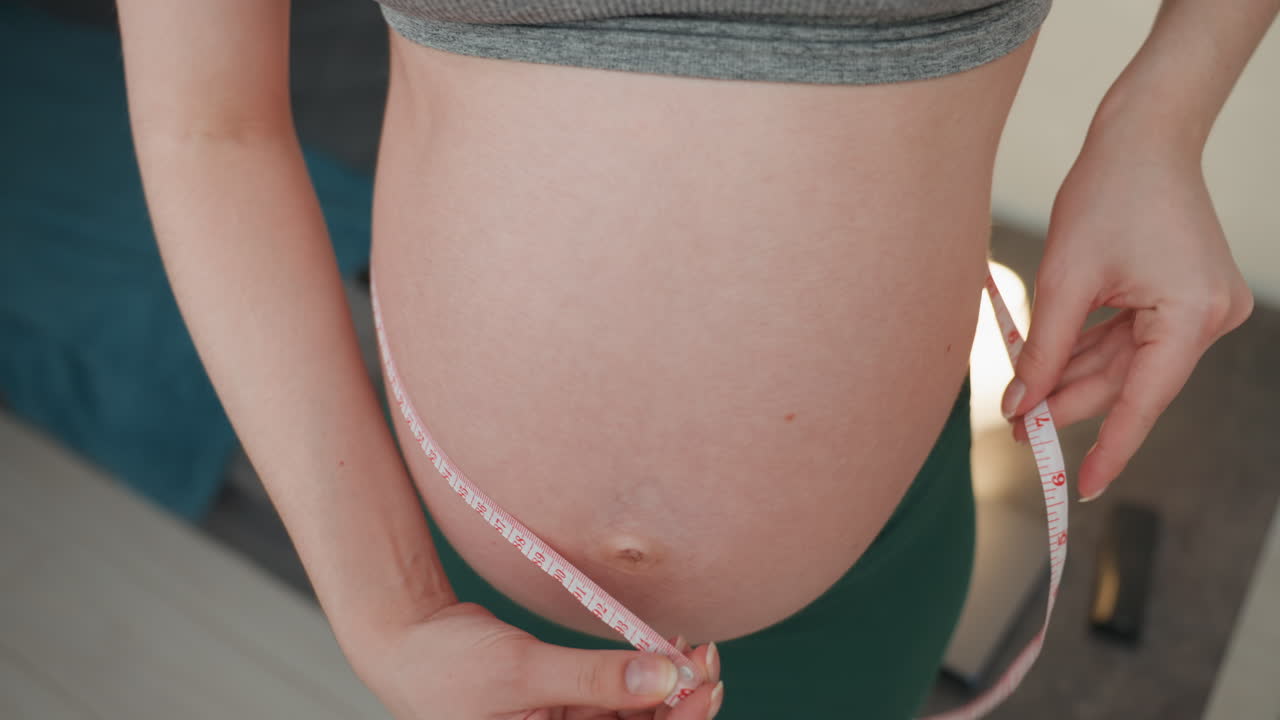 Close-up de mulher grávida medindo a barriga com fita métrica, luz natural suave, sutiã esportivo cinza, mãos ajustando a fita, fundo de quarto, doce expectativa, checagem pré-natal pessoal.