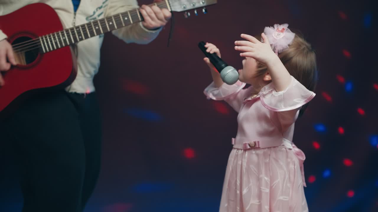 Llittle girl in vintage dress sings on stage, her father plays acoustic guitar