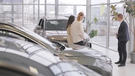 young car salesman showing to young couple new automobile at dealership salon.