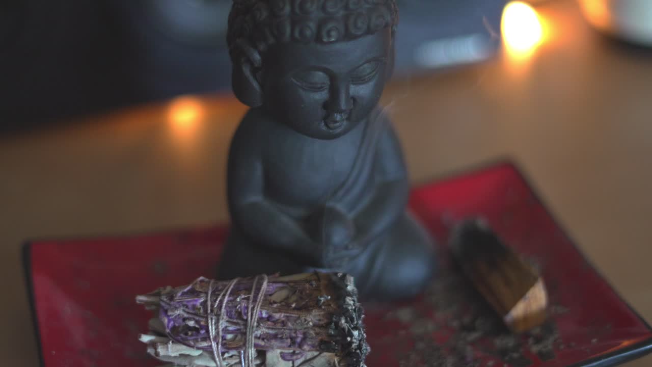 pequeña estatua de buda relajándose con salvia de lavanda humeante en llamas sentada en un plato asiático rectangular rojo en un estante de mesa en 4k 60fps