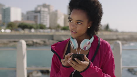 Retrato-De-Una-Atractiva-Mujer-Afroamericana-En-Ropa-Deportiva-Frente-A-La-Playa-Usando-Un-Teléfono-Inteligente